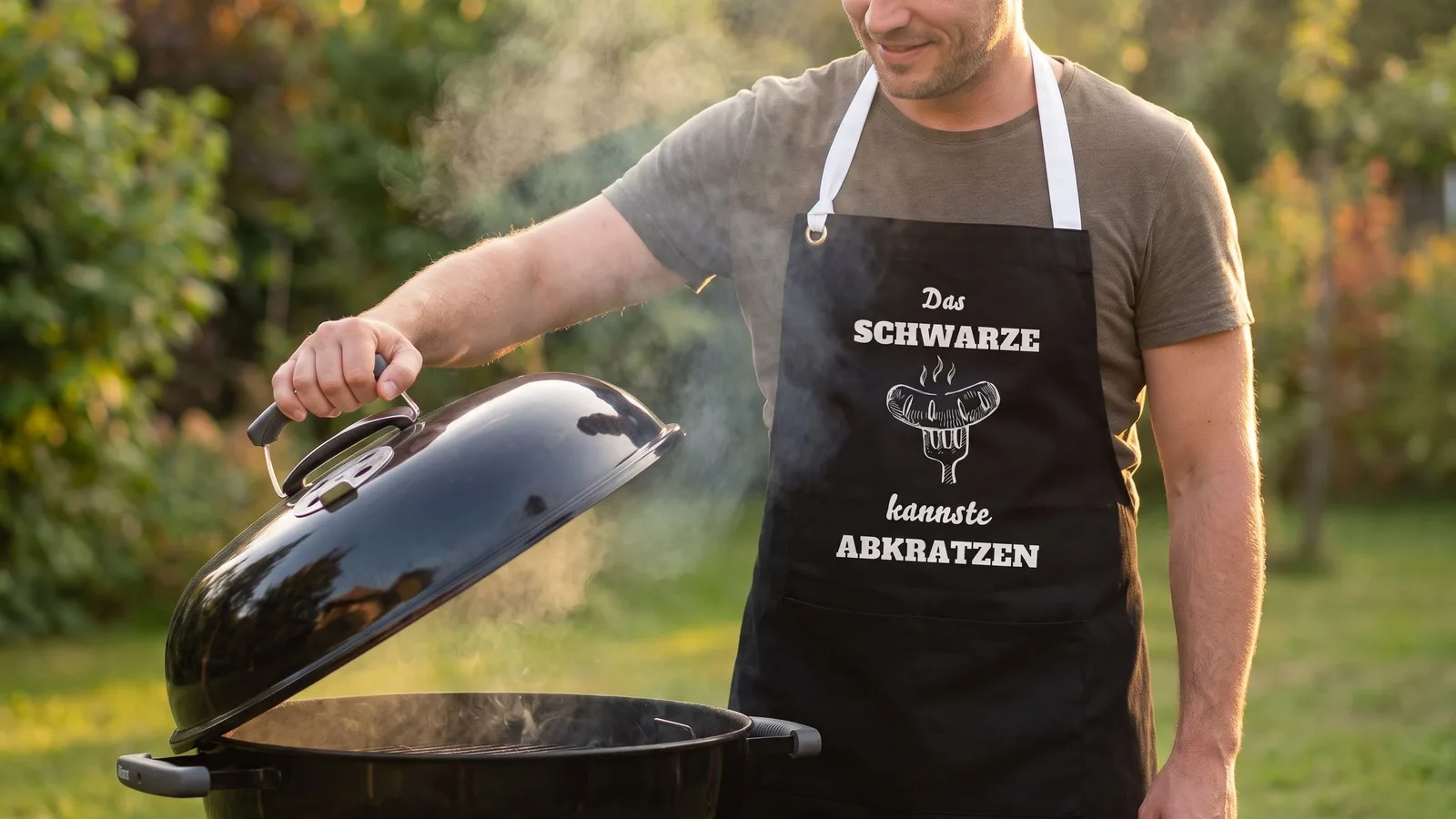 Mann hebt den Deckel eines Kugelgrills, schwarze Schürze mit dem Aufdruck „Das Schwarze kannste abkratzen“